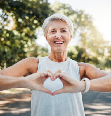 Fitness, happy and heart hands of old woman in nature after running for health, wellness and workout. Smile, motivation and peace with senior lady and sign for love, faith and training in nature